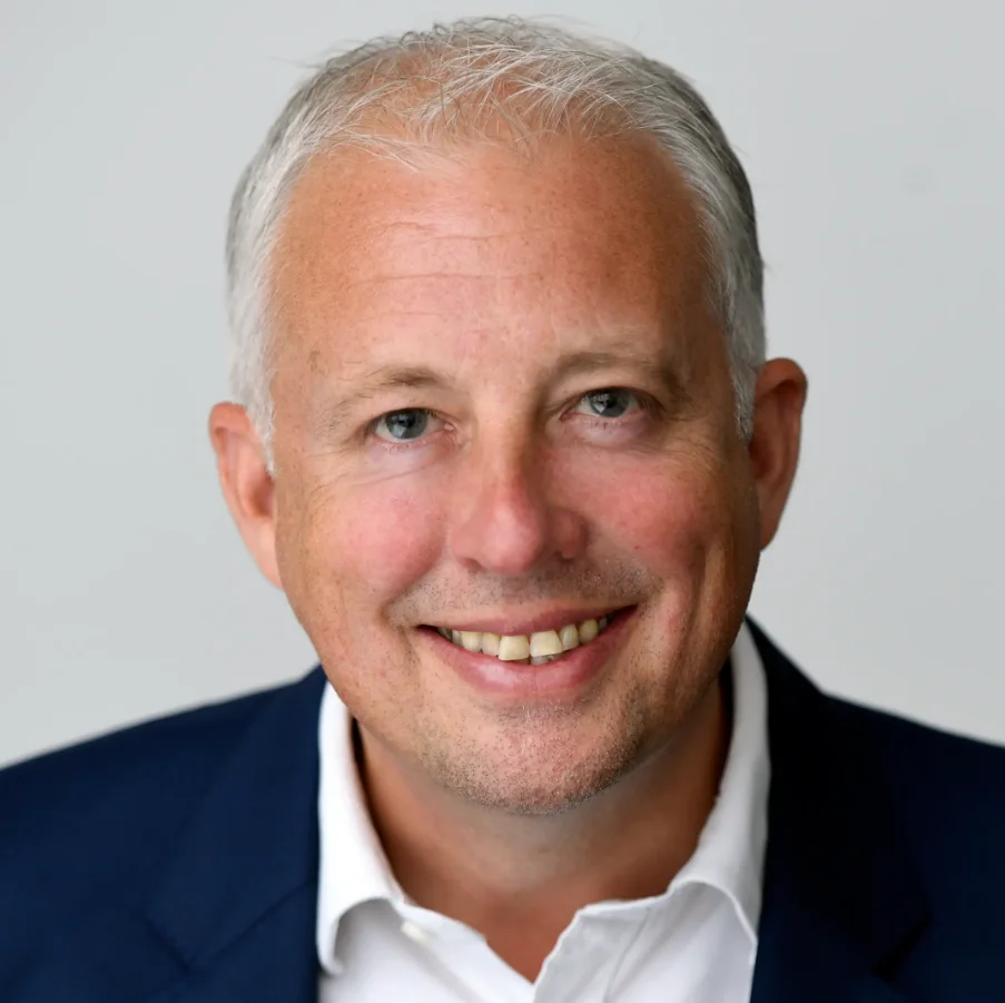 Chris Rolland smiles for a portrait wearing a navy jacket and white shirt against a grey background.
