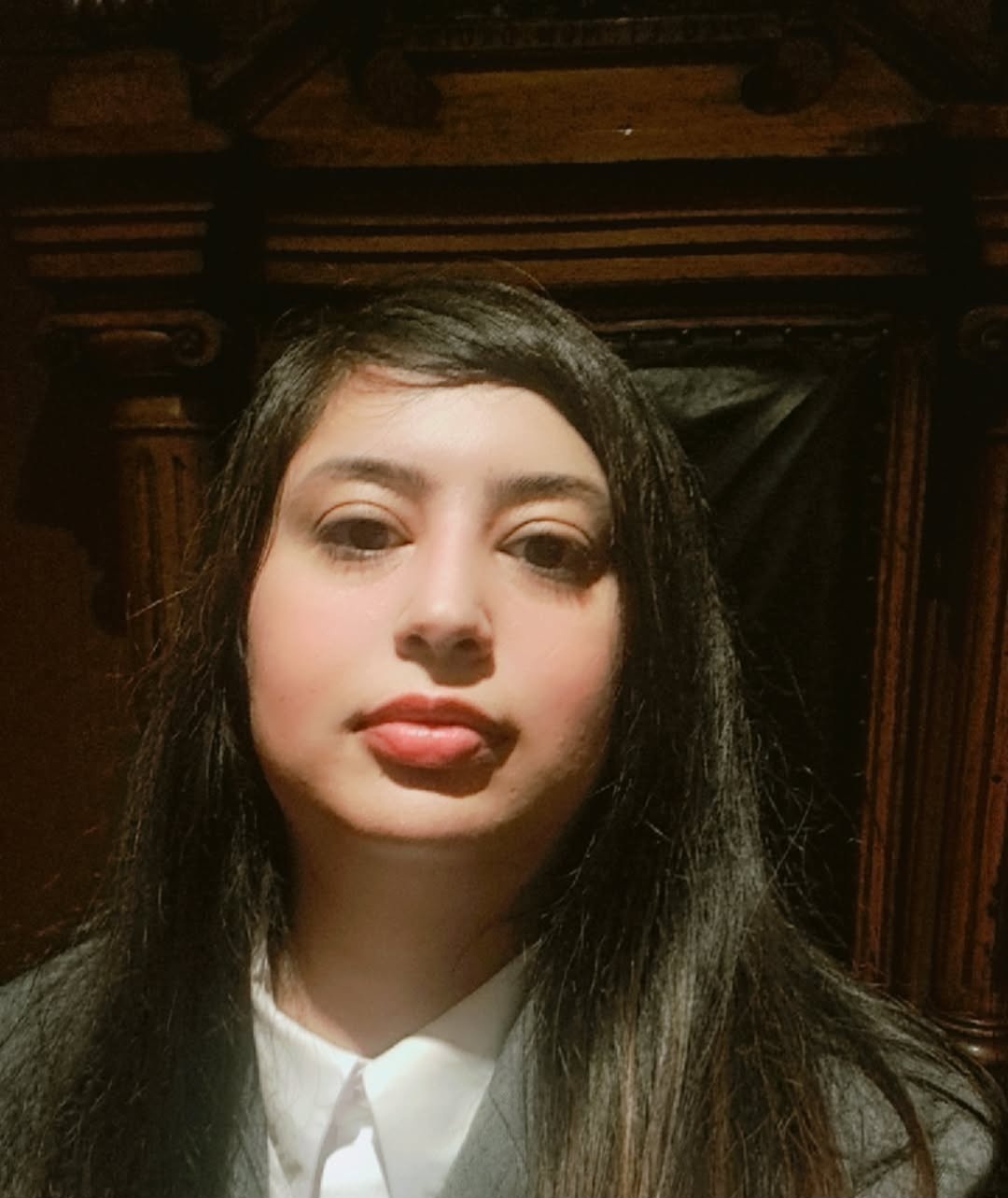 Dr Nesha Maqbool wearing a white collared shirt and grey jacket in front of dark wooden panelling.
