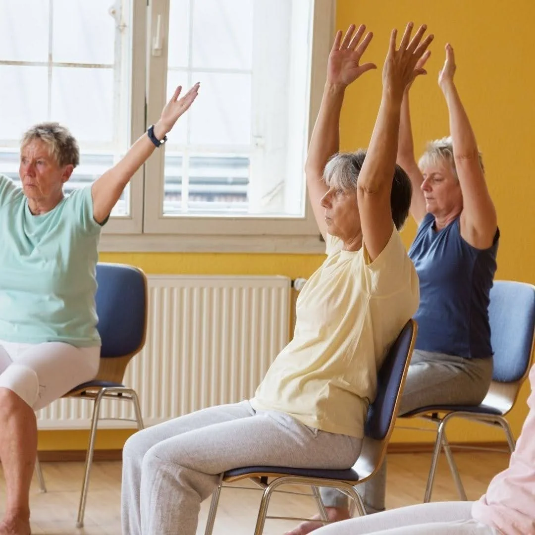 Three older women seated on chairs in a bright yellow room raise their arms overhead during a gentle chair yoga session.