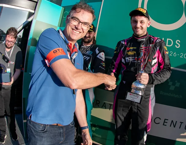 Andy Williams in a blue polo shirt shakes hands with racing driver Aaron Cooke, who is wearing his race suit and holding his GT Cup Championship trophy at Brands Hatch.