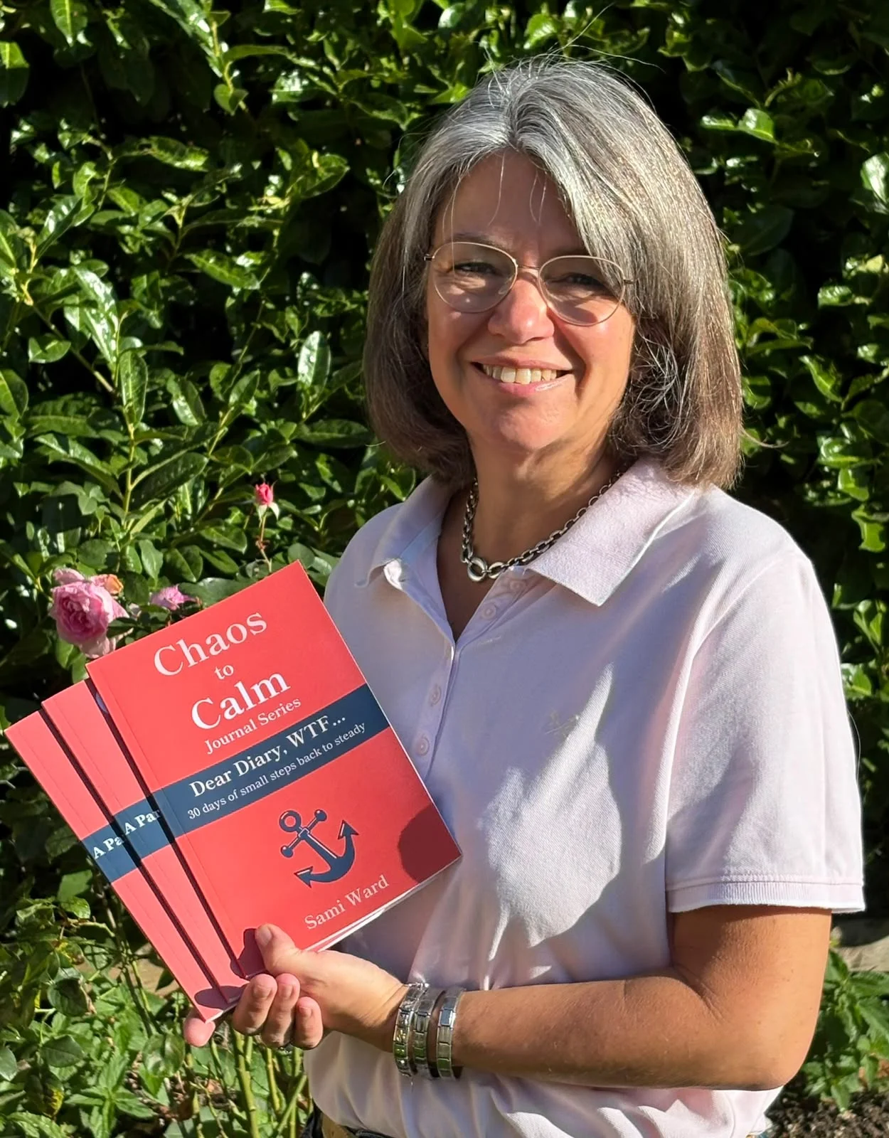 Sami Ward smiles outdoors in front of green foliage while holding three red Chaos to Calm books, including the journal Dear Diary, WTF...